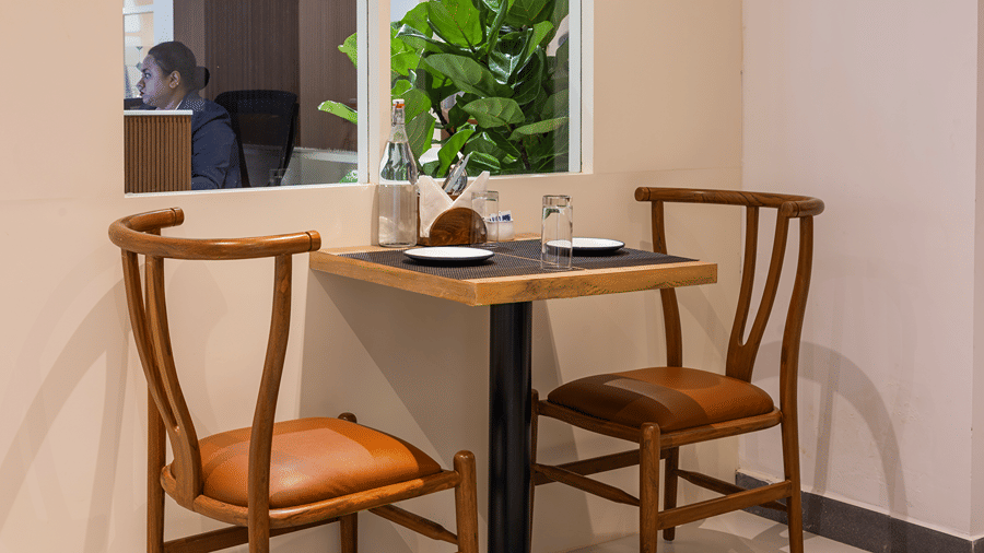 Table for 2 with wooden chairs, place settings and a window looking onto the reception at M-Salt restaurant at Monday Hotels & Suites Madhapur, Hyderabad.