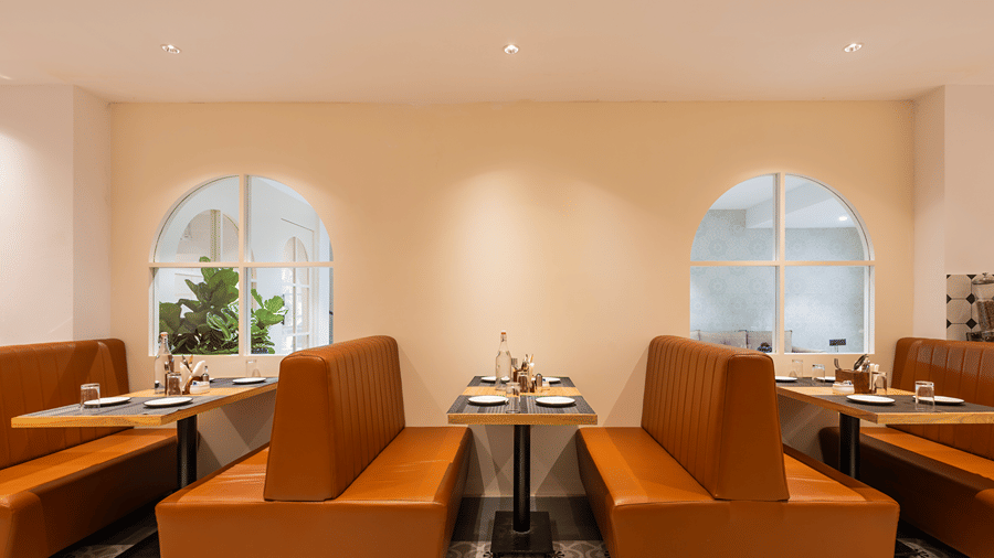 Tan leather booths with set tables, glasses and cutlery holders in front of 2 arched windows at M-Salt restaurant at Monday Hotels & Suites Madhapur, Hyderabad.