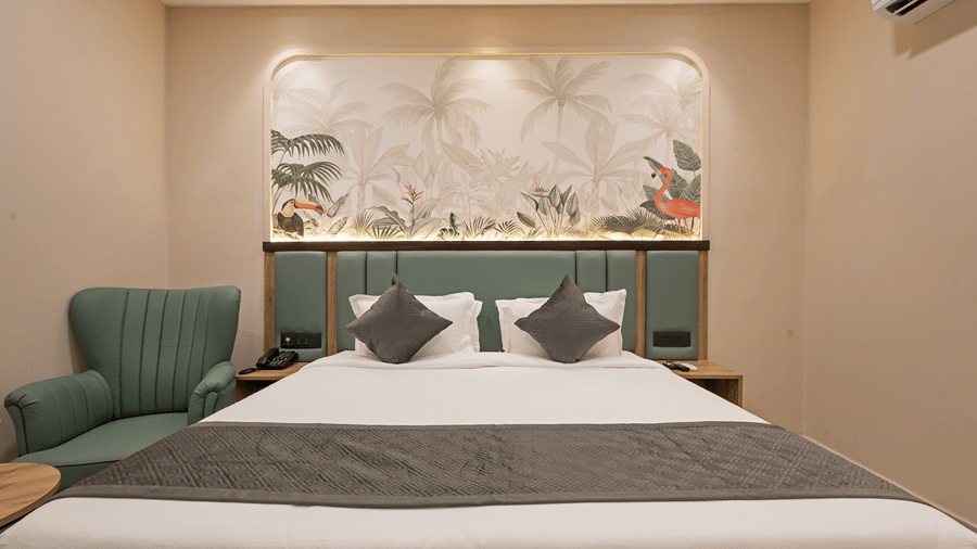 King bed with grey cushions below a toucan and flamingo mural, green armchair and bedside phone in a Superior room at Monday Hotels & Suites Madhapur, Hyderabad.
