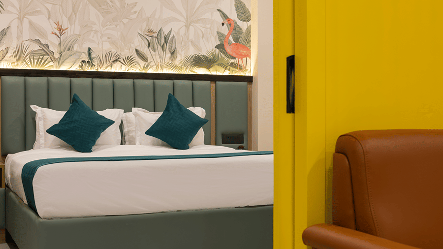 Bed with teal cushions under a flamingo mural beside the yellow sliding door and tan armchair in a Studio Suite at Monday Hotels & Suites Madhapur, Hyderabad.
