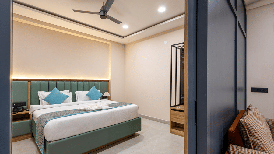 A bedroom with a king size bed made with white linens and blue pillows is viewed beside a dark partition wall at Monday Hotels & Suites Madhapur Hyderabad.
