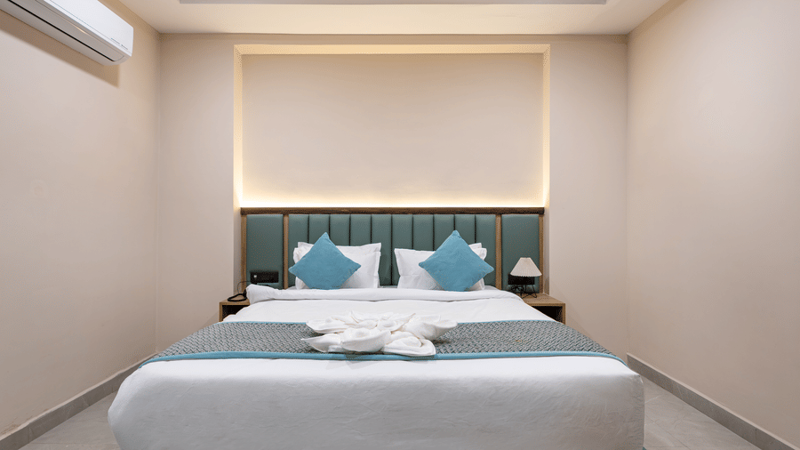 A king bed with white linens and blue accent pillows, 2 side tables, and an air-conditioner in a guest room at Monday Hotels & Suites Madhapur Hyderabad.