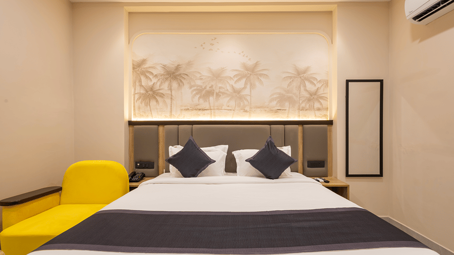 King bed with navy cushions below a sepia palm mural, yellow armchair and wall mirror in a Premium room at Monday Hotels & Suites Madhapur, Hyderabad.