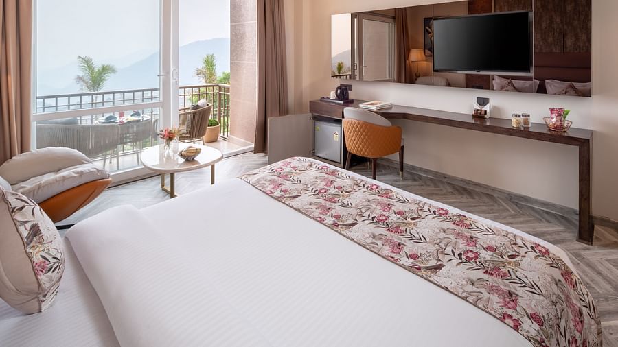 an image of a bedroom with balcony from where sunlight is flooding the room which has a flat TV screen,  a narrow desk below it paired with a chair.