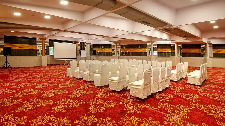 A spacious banquet hall with a patterned red carpet, rows of white-covered chairs facing a projector screen, and a reflective ceiling creating an elegant atmosphere.