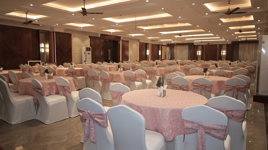 Spacious hotel banquet hall with round tables, white chairs with pink sashes, and elegant ceiling lighting set for events.