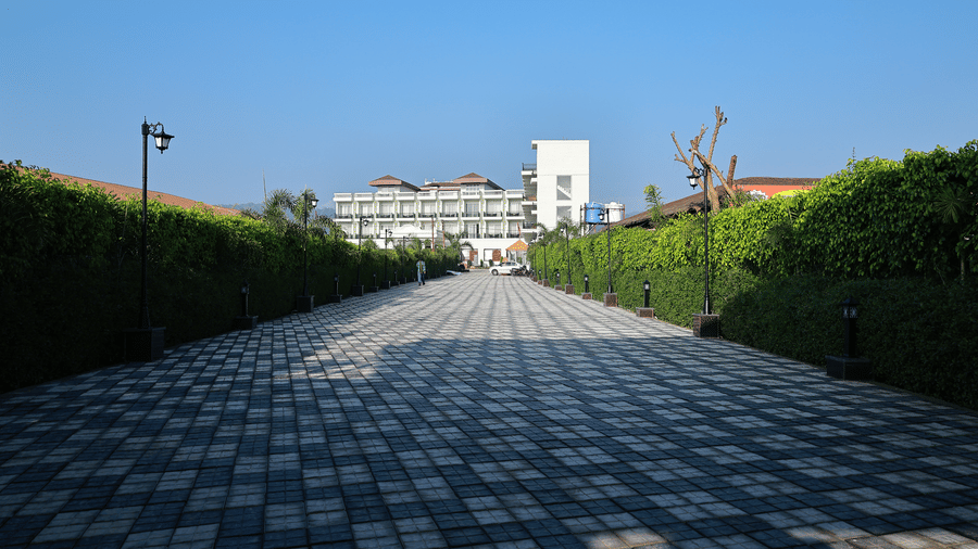 A long, patterned stone driveway lined with tall green hedges and street lamps leading toward the white resort building.