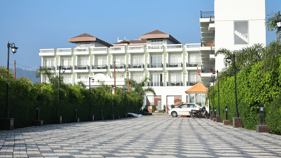 Long paved walkway lined with green hedges leading toward the main white hotel building and parking area.
