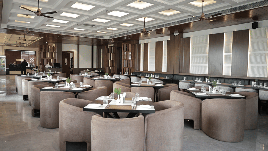 Modern hotel dining hall featuring multiple round cushioned chairs, well-set tables, warm lighting, and contemporary ceiling design.