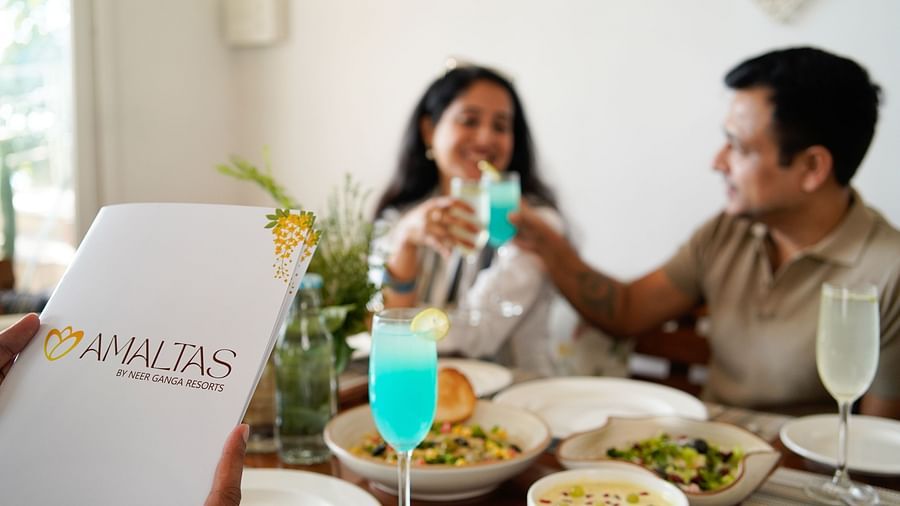 A close-up of the Amaltas menu with two people in the background cheering with glasses, surrounded by a variety of food and drinks at Neer Ganga.