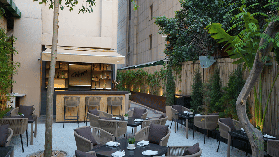 A chic outdoor cafe courtyard with dining sets leading toward an exterior bar counter under a tree.