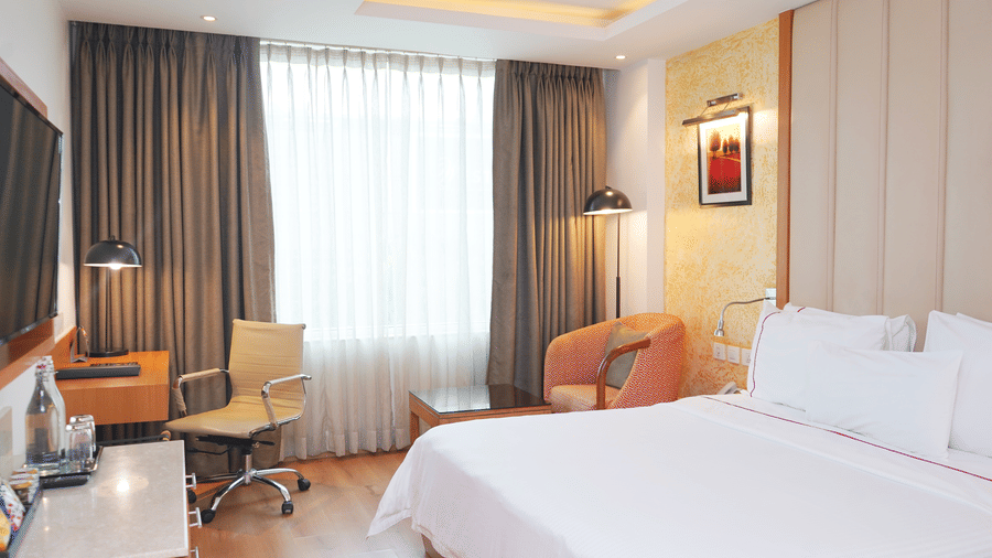 A bright hotel suite with a white bed, an orange armchair by the window, dark curtains, a work desk, and a wall-mounted TV.