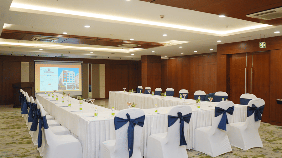 A conference room arranged in a u-shape configuration with white chairs wrapped in dark blue sashes.