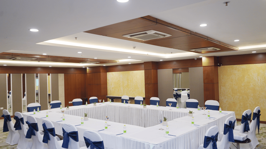 A large meeting room arranged in a hollow square layout with tables covered in white tablecloths and chairs with blue bows.