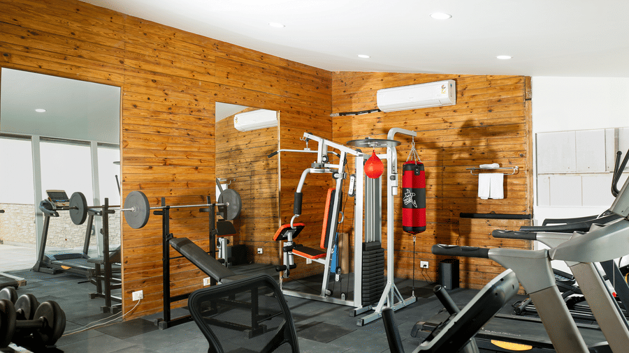 A well-equipped fitness room with cardio equipment, weight lifting machines, and an air conditioner mounted on a wood wall.