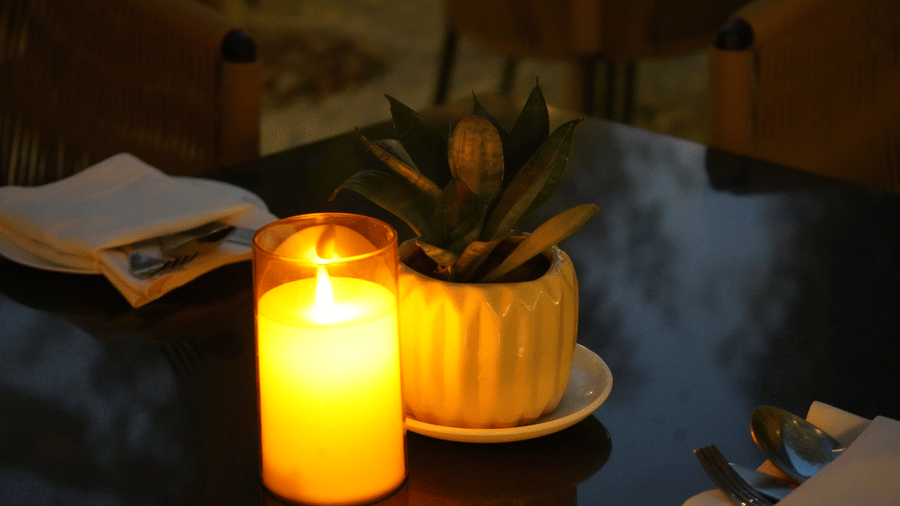 Close-up of a glowing yellow candle in a glass cylinder next to a potted succulent on a dark restaurant table.