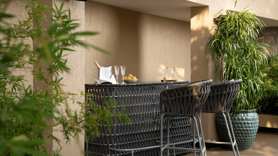 A stylish outdoor bar counter with tall wicker stools surrounded by potted plants and green foliage.