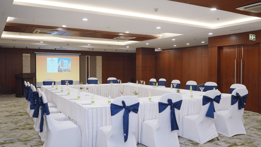 A meeting room arranged in a square layout with white tablecloths, chairs with dark blue bows, and a projector screen.