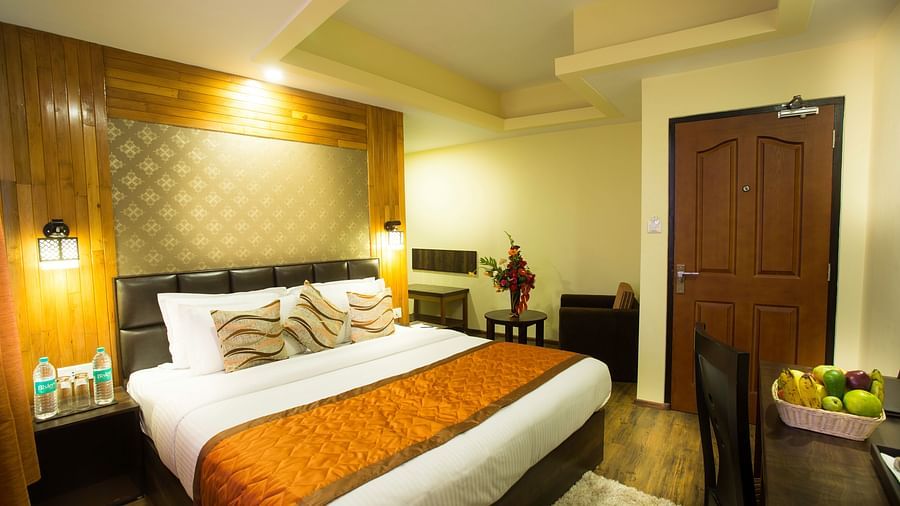 A room at Hotel New Orchid in Gangtok contains a bed with pillows, bedside tables with lamps and water bottles, a desk with a chair and fruit bowl, and a do