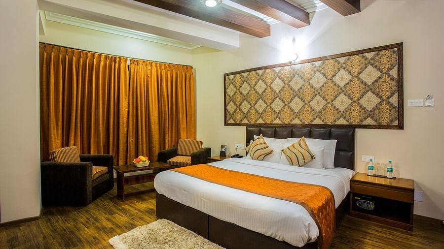 A room at New Orchid Lords Inn Gangtok contains a bed with pillows, bedside tables with lamps and water bottles, chairs, a coffee table with fruit, curtains, and a rug.