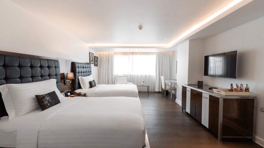 One of the best Bangkok places to stay, featuring two large beds, white linens, dark headboards, a flat-screen TV, minibar, and a window letting in natural light at the far end of the room at Night Hotel Bangkok.