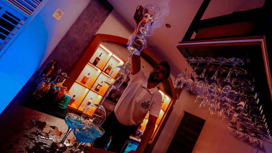 Low angle shot of a bartender pouring a drink into a glass, with rows of glasses and bottles visible in the background - Nirantara Resort & Spa.