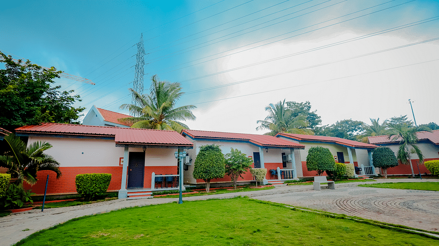 Exterior view of cottages with red roofs and green lawns at Nirantara Resort & Spa, Bengaluru, under a partly cloudy sky.