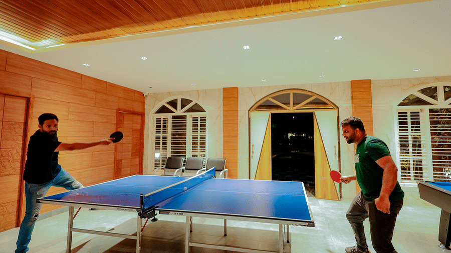 Two people play ping pong in a room with brick walls, large windows, and a high ceiling with a decorative light fixture - Nirantara Resort & Spa.