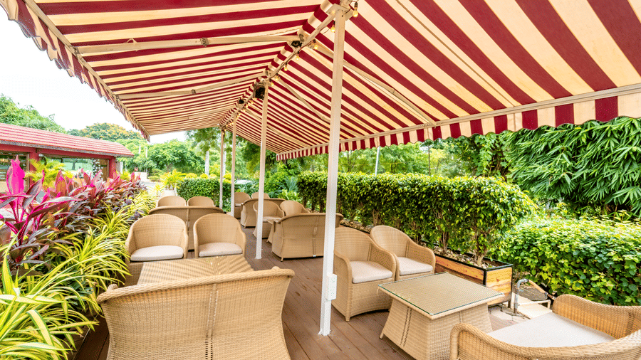 Alternative view of Café Latte at Noor-Us-Sabah Palace, Bhopal, featuring wicker lounge seating arranged on a wooden deck beneath a large awning.