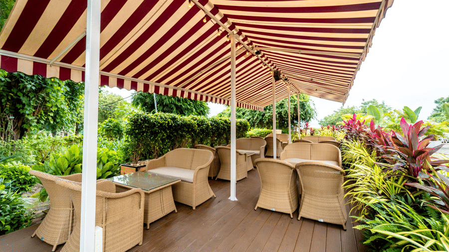 The outdoor seating area surrounded with green plants at Café Latte, Noor-Us-Sabah Palace, Bhopal, with wicker seating on a wooden deck under a red and white striped awning.