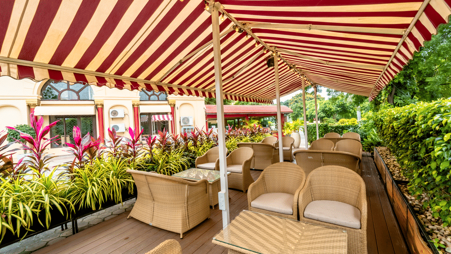 Wicker chairs and glass-topped tables at Café Latte, Noor-Us-Sabah Palace, Bhopal, situated on an outdoor patio bordered by tropical plants.