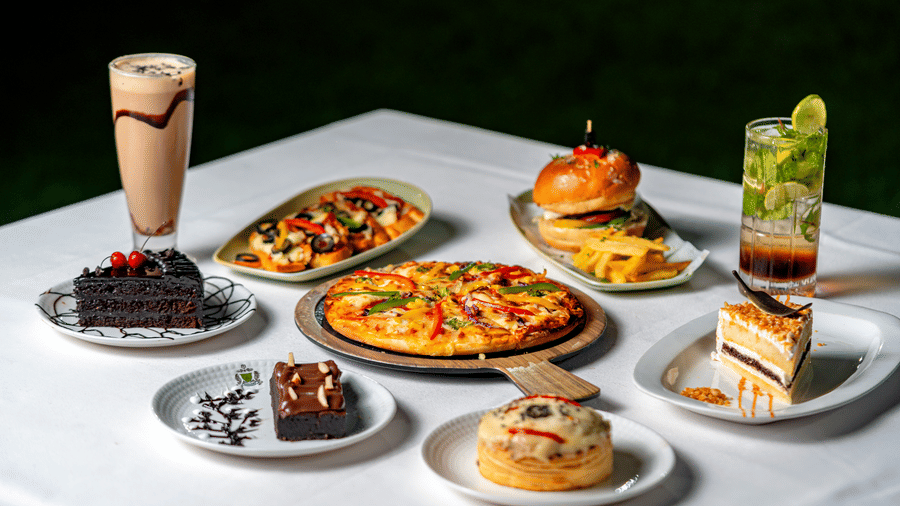 A spread of pizza, burgers, and desserts at Café Latte, part of the Noor-Us-Sabah Palace, Bhopal, served on a white tablecloth with various drinks.