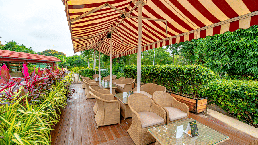 Outdoor seating area with a wet wooden deck at Café Latte, found at Noor-Us-Sabah Palace, Bhopal, featuring wicker armchairs and glass tables under a striped awning.