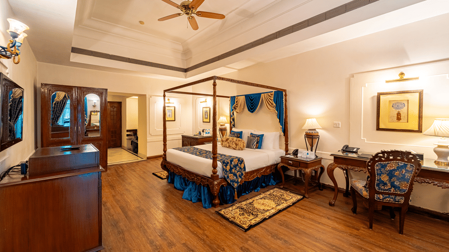 A wide-angle shot of the bedroom of the Crown Club Suite at Noor-Us-Sabah Palace, featuring a grand four-poster bed with royal blue accents, a wooden desk, and polished floors.