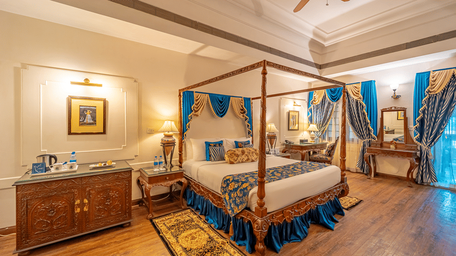 Full view of the elegant bedroom of the Crown Club Suite at Noor-Us-Sabah Palace, Bhopal, showing the four-poster bed and traditional wooden sideboard.