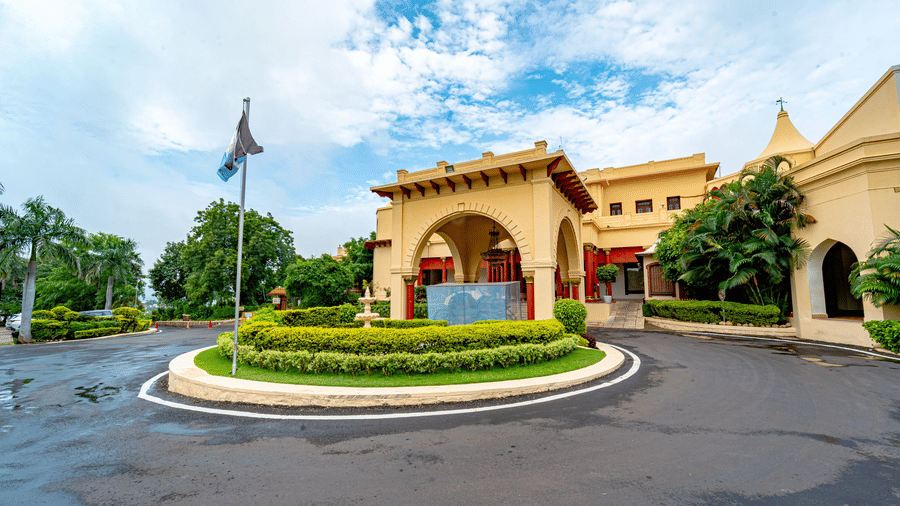 The grand main entrance of Noor-Us-Sabah Palace, Bhopal, featuring a circular driveway, manicured green hedges, and traditional yellow heritage architecture under a blue sky.