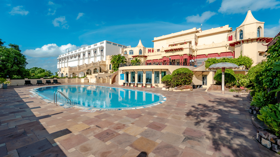 A wide view of the large outdoor swimming pool at Noor-Us-Sabah Palace, Bhopal, set against the backdrop of the heritage hotel's white and yellow facade under the vast blue sky.