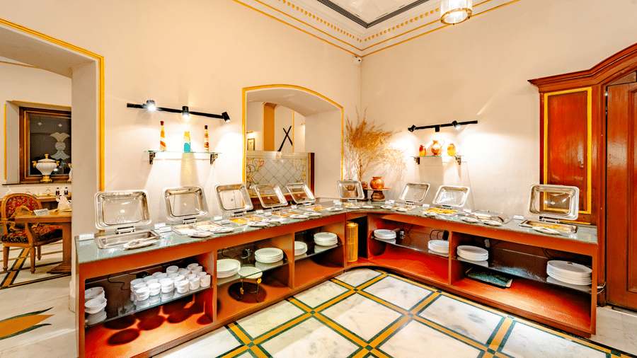 A long buffet counter at Marble Arch, found at Noor-Us-Sabah Palace, Bhopal, featuring silver serving dishes, wooden cabinetry, and marble flooring.