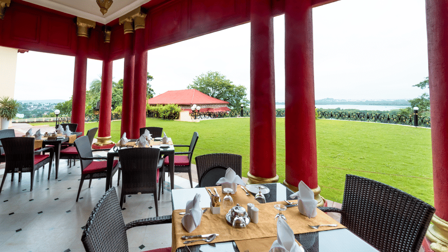 A wide-angle shot of the outdoor seating area under a roof at the Marble Arch restaurant with lake and lawn views at Noor-Us-Sabah Palace, Bhopal.