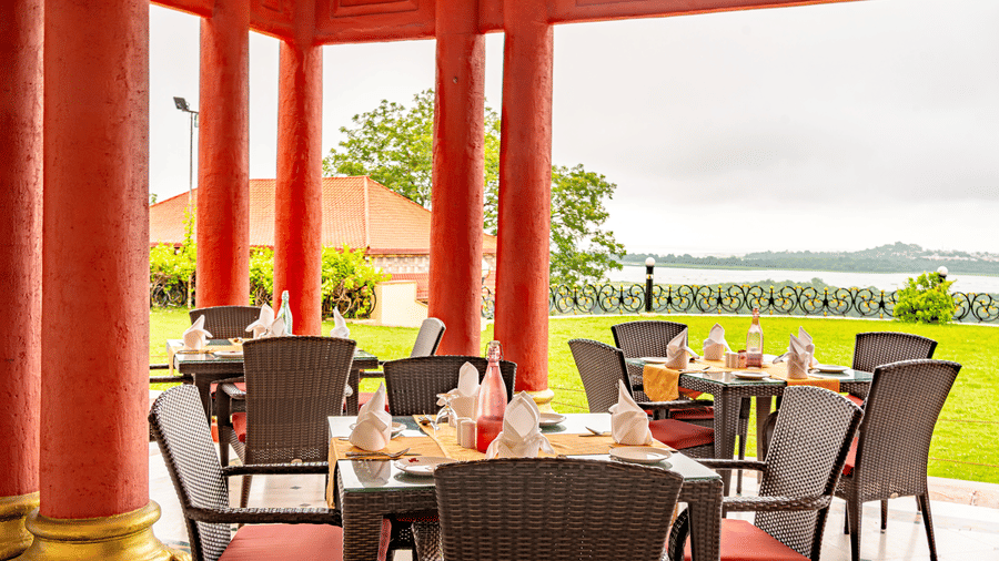 The outdoor dining area of the Marble Arch multi-cuisine restaurant in Bhopal at the Noor-Us-Sabah Palace overlooking the lush green lawn and the lake under a cloudy sky in the background.