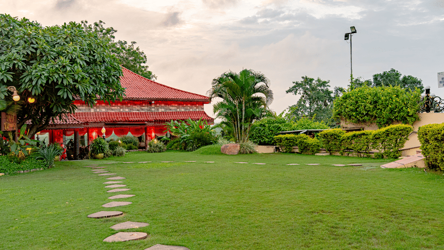 An exterior view of Ming Dynasty at Noor-Us-Sabah Palace, Bhopal, showing a stone pathway leading across a manicured lawn toward the traditional red-roofed restaurant.