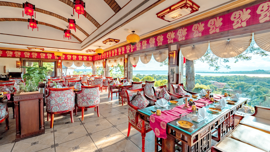 The interior of Ming Dynasty at Noor-Us-Sabah Palace, Bhopal, showing an elegantly set dining area with red patterned chairs and large windows that offer a panoramic view of the lake and greenery.