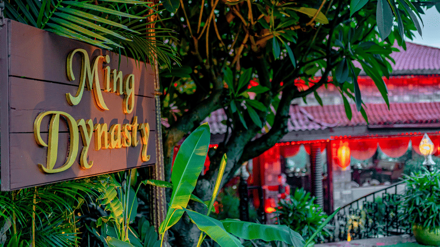 The glowing entrance sign of Ming Dynasty, found at Noor-Us-Sabah Palace, Bhopal, surrounded by tropical foliage and the warm evening lights of the restaurant.