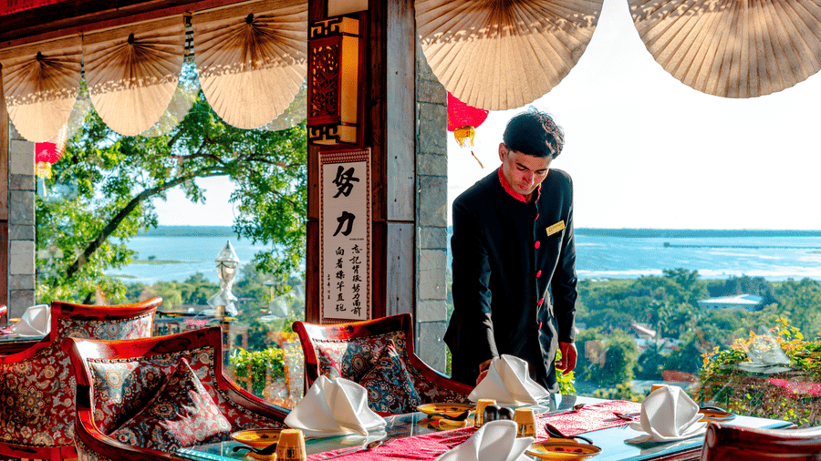 A staff member preparing an indoor table for guests at Ming Dynasty, located within Noor-Us-Sabah Palace, Bhopal, featuring large windows that provide a panoramic view of the lake.