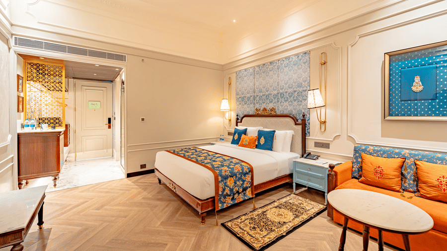 A wide view of the Premium Club Room at Noor-Us-Sabah Palace, Bhopal, featuring a large bed with turquoise and orange accents, light wood herringbone floors, and an orange sofa.