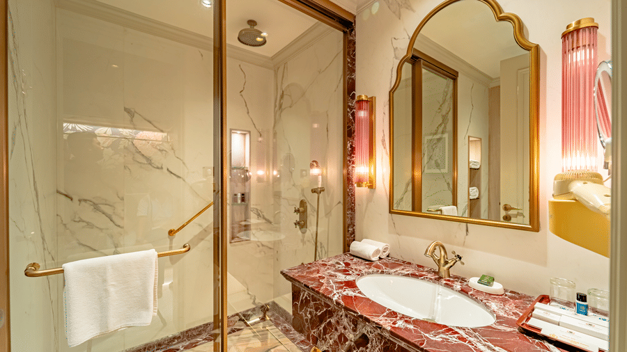 The luxurious bathroom of the Premium Club Room at Noor-Us-Sabah Palace, Bhopal, featuring a red marble vanity, a gold-framed arch mirror, and a glass-enclosed walk-in shower.