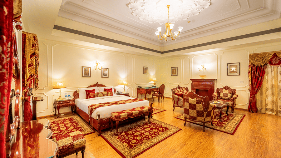 A wide view of the Presidential Suite, featuring a large bed with red and gold checkered linens, a separate seating area with matching armchairs, polished wooden floors, and an ornate white ceiling with a central chandelier at Norr-Us-Sabah Palace, Bhopal.