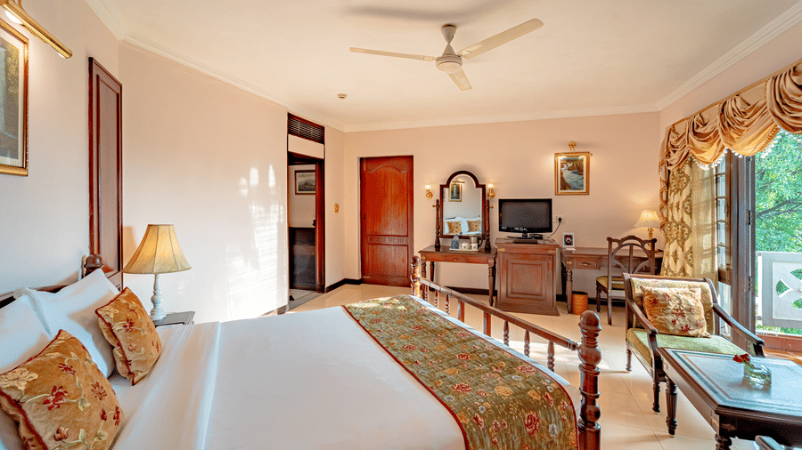A bright Standard Room at Noor-Us-Sabah Palace, Bhopal, featuring a classic wooden bed with floral linens, a study desk, and a large window with gold curtains.