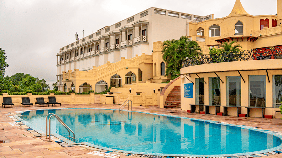 A swimming pool in the foreground with a beige-coloured facade of Noor-Us-Sabah Palace, Bhopal, against a cloudy sky in the background.