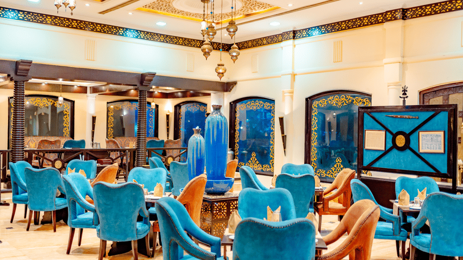 The main dining area of Za-aiqa, located within Noor-Us-Sabah Palace, Bhopal, featuring vibrant teal velvet chairs, arched wall alcoves, and a spacious, elegant layout.